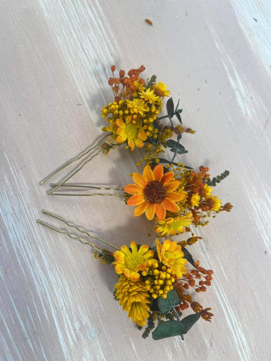Three yellow and orange flower hair pins with green leaves and brown stems, arranged on a light gray wooden background.