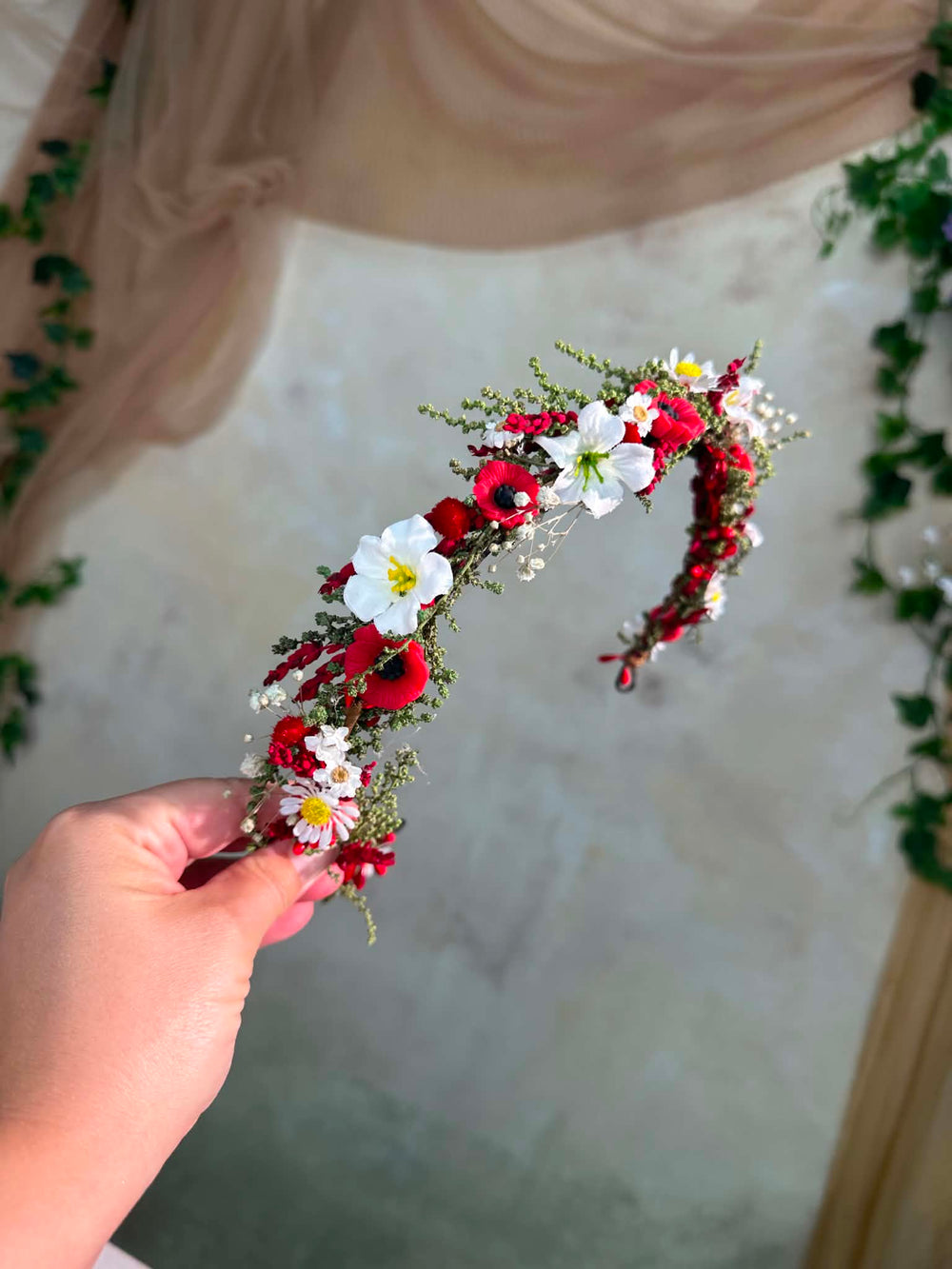Hand holding a floral headband with red, white, and green flowers against a neutral background.