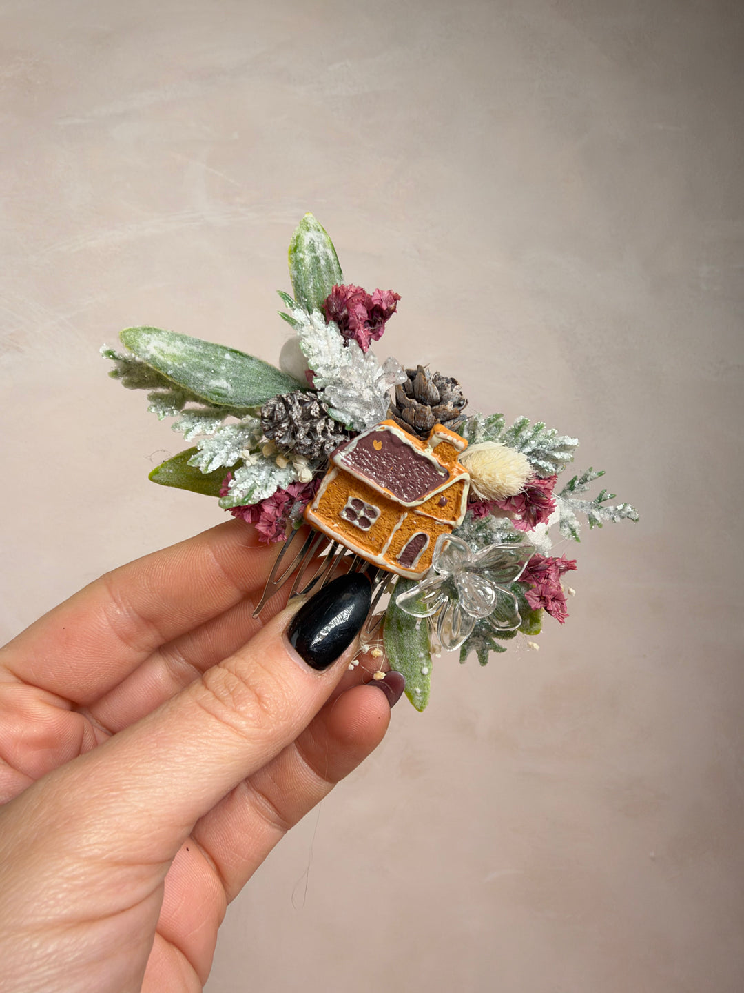 A hand with black nail polish holds a decorative floral hair accessory featuring a small gingerbread house, frosted greenery, and dusty pink berries against a plain beige background.