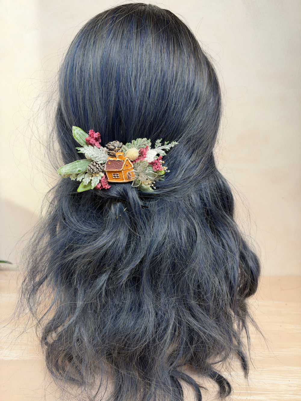 A long, wavy dark gray wig with a small decorative hair accessory featuring green leaves, pink flowers, and a tiny orange house, shown from the back on a mannequin head against a light beige background.