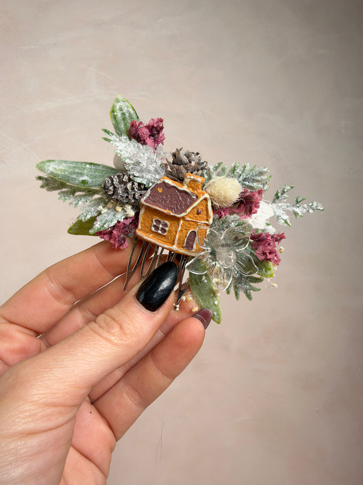 A hand with black nail polish holds a small decorative hair accessory featuring a miniature gingerbread house, frosted green leaves, dusty pink berries, and silver glitter accents against a plain beige background.