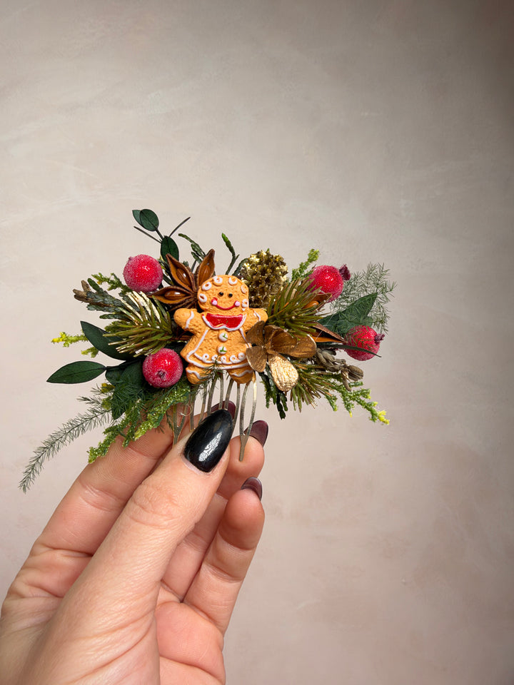 A hand with black glitter nail polish holds a small Christmas hair accessory featuring a gingerbread man figure surrounded by green pine branches, brown foliage, and bright red berries, set against a plain beige background.