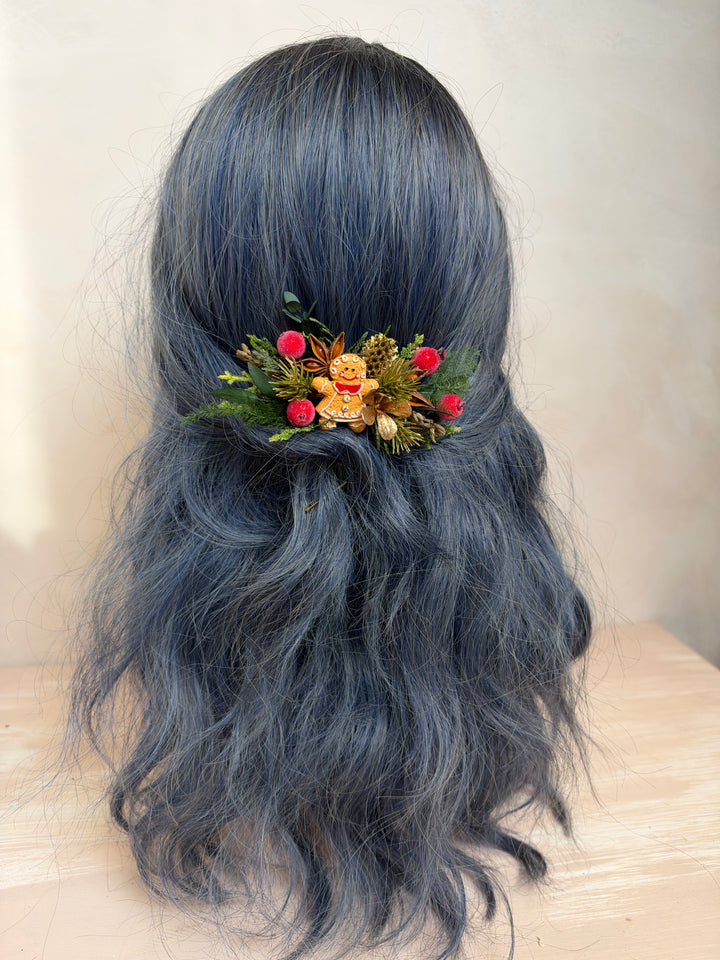 A long, wavy dark blue-gray wig displayed on a mannequin head, styled with a small autumn floral hair accessory featuring red berries, green leaves, and a tiny gingerbread figure.