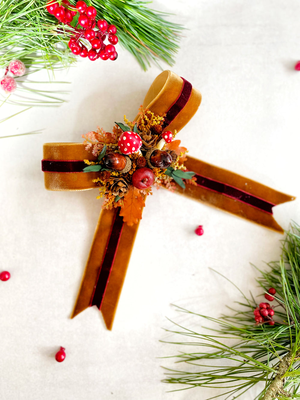 A decorative Christmas ornament shaped like a ribbon cross, made from brown velvet ribbon with dark red stripes, adorned with red mushrooms, pinecones, and small autumn leaves, photographed on a light textured background with pine branches and red berries as styling props.