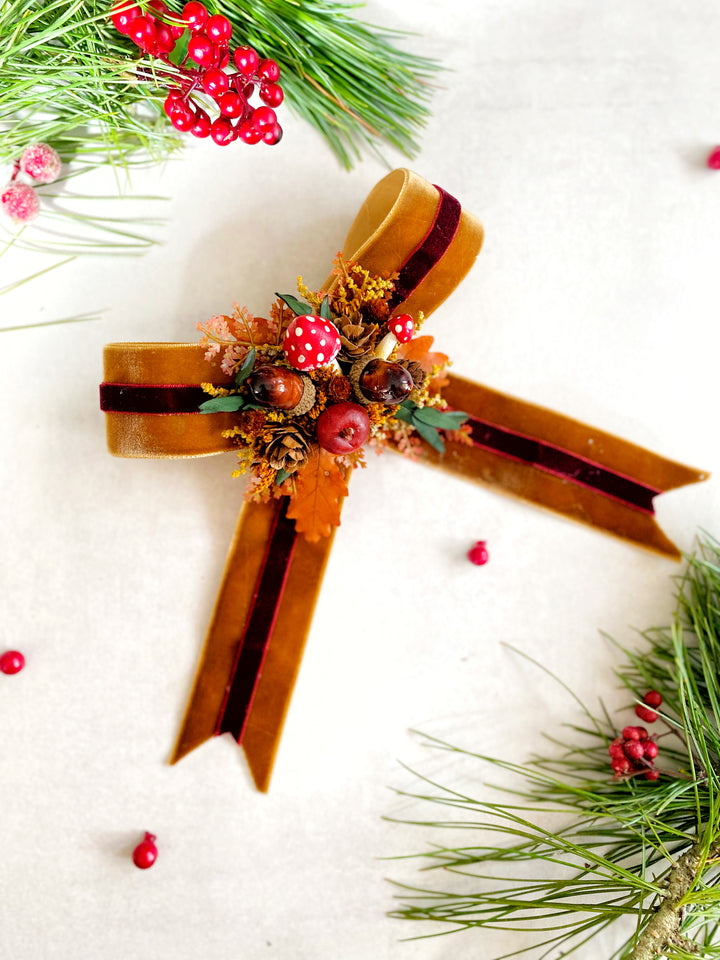 A decorative Christmas ornament shaped like a ribbon cross, made from brown velvet ribbon with dark red stripes, adorned with red mushrooms, pinecones, and small autumn leaves, photographed on a light textured background with pine branches and red berries as styling props.