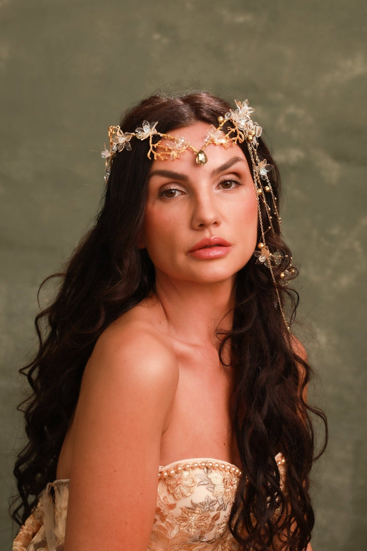 A woman with long, wavy dark brown hair wears an ornate gold and white floral headpiece with dangling chains and a central jewel, posed against a textured olive green studio background.