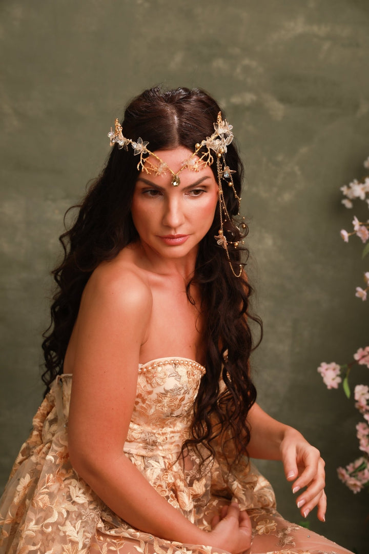 A woman with long dark wavy hair wears an ornate gold and white floral headpiece with dangling earrings and a strapless champagne-gold patterned gown, posed against a textured olive-green backdrop with soft pink flowers at the side.