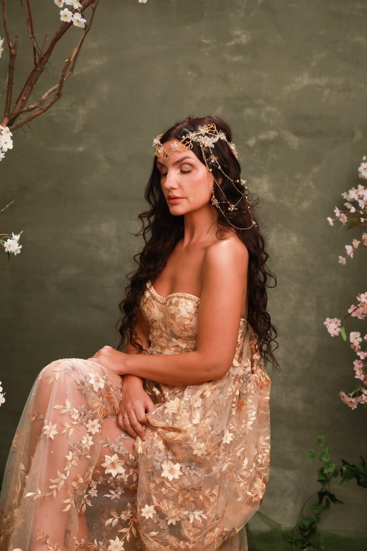 A woman seated in a sheer champagne-gold embroidered gown with a matching floral headpiece, posed against a textured olive-green backdrop with soft pink blossoms at the edges.
