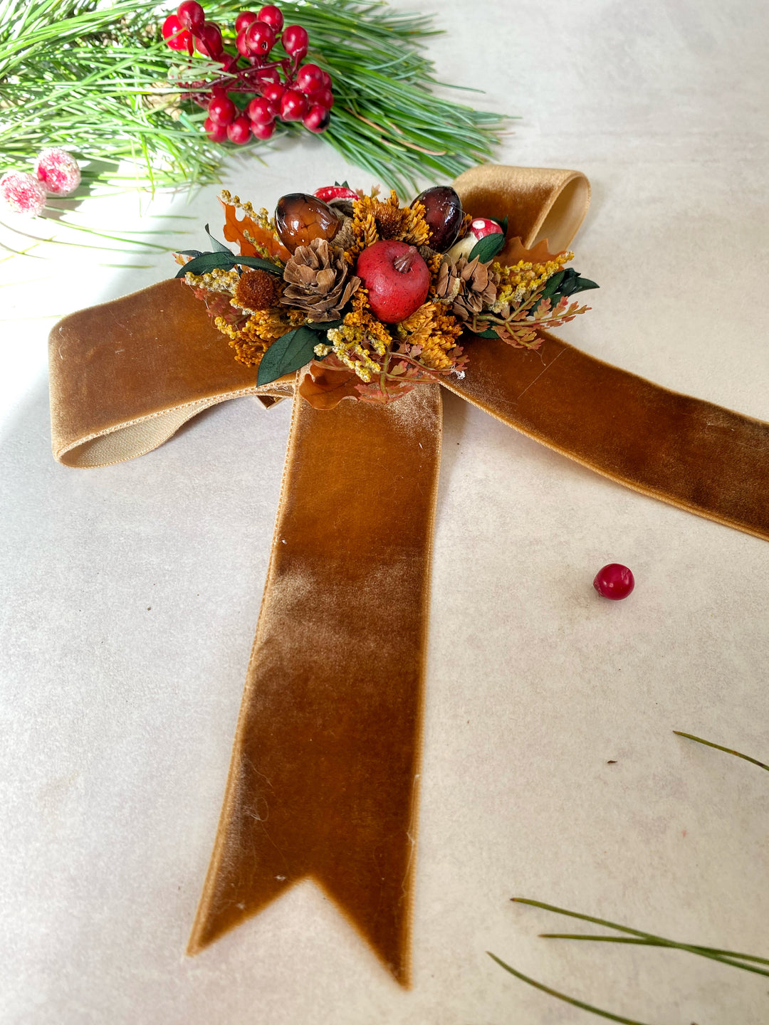 A decorative autumn-themed ribbon bow made of wide tan velvet, with a center cluster of dried orange leaves, pinecones, acorns, small red berries, and green foliage, photographed on a light neutral surface with holiday greenery in the corners.
