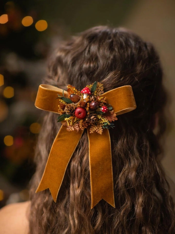 A dark brown wavy hair accessory featuring a large mustard yellow velvet bow with a festive dried botanical cluster in the center, shown worn on a person's head against a warm, blurred holiday background.