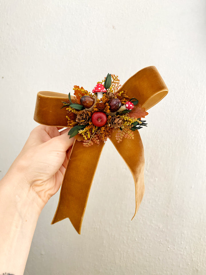 A hand holds a large tan velvet ribbon bow decorated with dried autumn foliage, acorns, red mushrooms, and berries against a plain off-white wall.