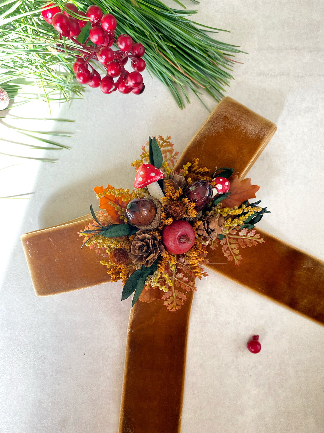 A rustic wooden hair bow decorated with autumn and Christmas elements, including pinecones, acorns, red mushrooms, berries, and a small red apple, photographed from above on a light neutral background with holiday greenery in the corner.