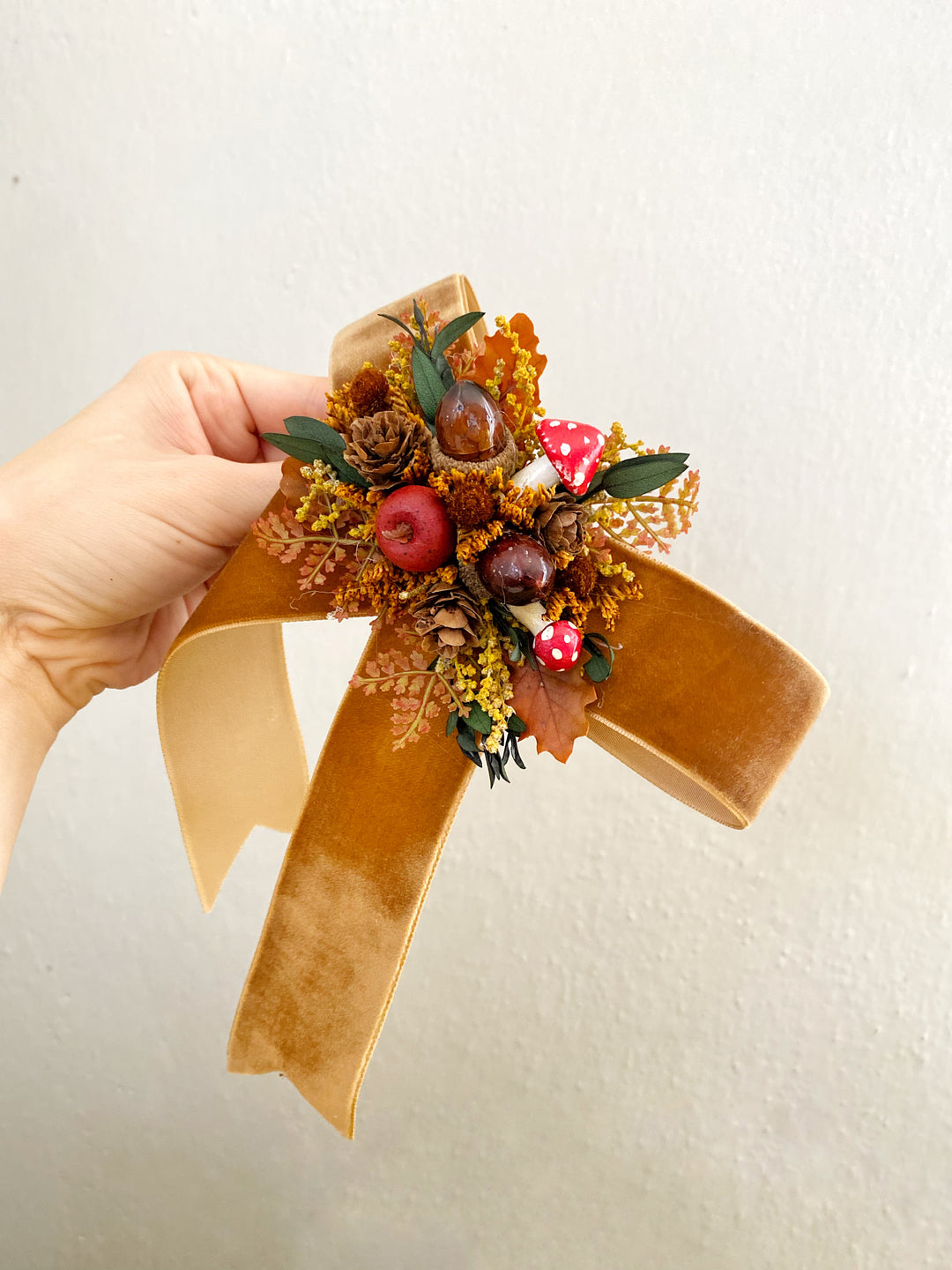 A hand holding a decorative autumn-themed hair bow accessory with a tan velvet ribbon, dried orange and brown leaves, pinecones, acorns, small red berries, and green leaves against a plain off-white wall.