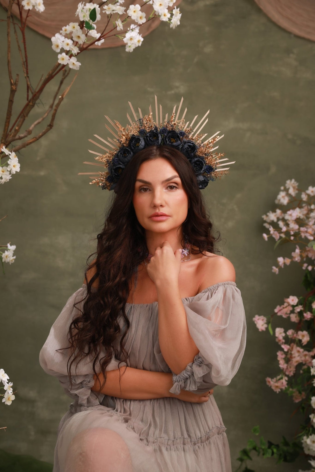 A woman wearing a dramatic gold spiked crown and a dark floral headpiece, posed in a sheer light gray off-the-shoulder dress against a textured olive-green backdrop with cherry blossom branches and pink flowers.