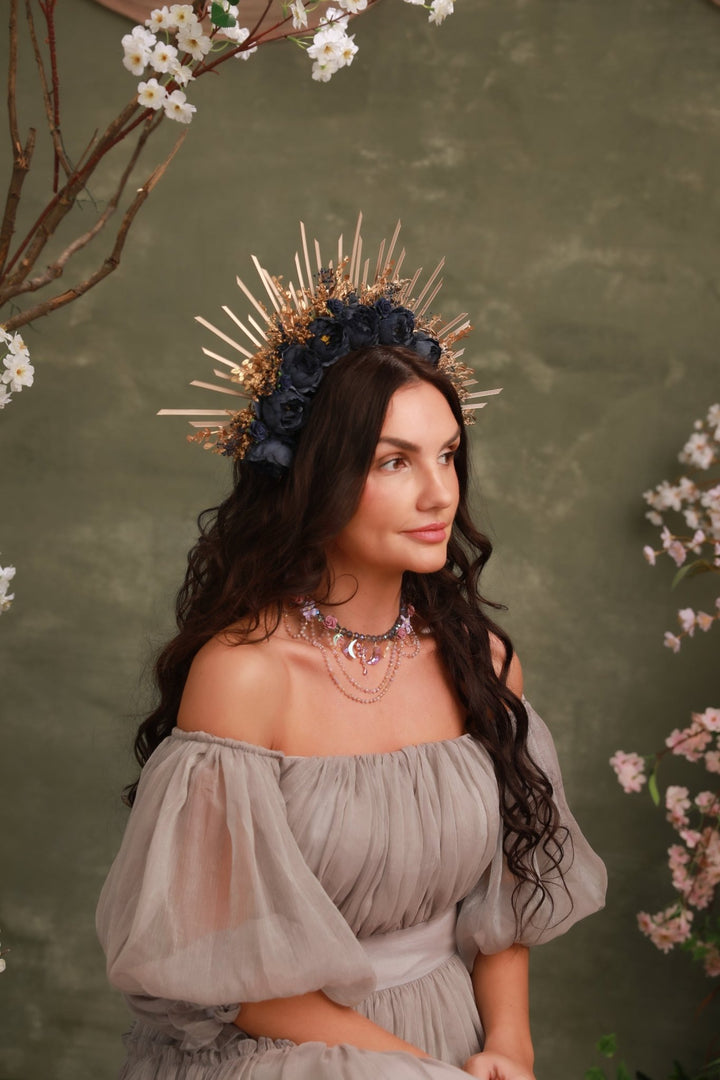 A woman wearing a dramatic gold and navy floral crown sits against a textured olive green backdrop with cherry blossom branches framing the scene. She wears a sheer off-the-shoulder taupe-gray gown and layered necklaces.