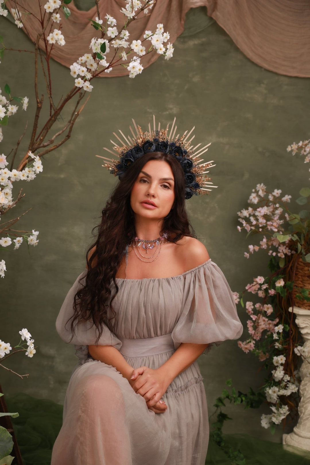 A woman seated against a textured olive-green backdrop with floral arrangements, wearing a sheer off-the-shoulder taupe dress and a metallic gold spiked crown.