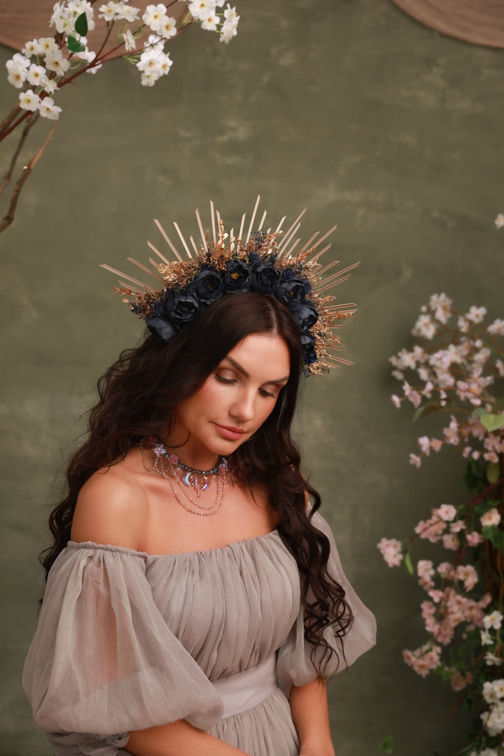 A woman wearing a decorative gold and navy floral crown with spiky gold accents, paired with a sheer off-the-shoulder taupe dress, posed against a textured olive green background with cherry blossom branches.