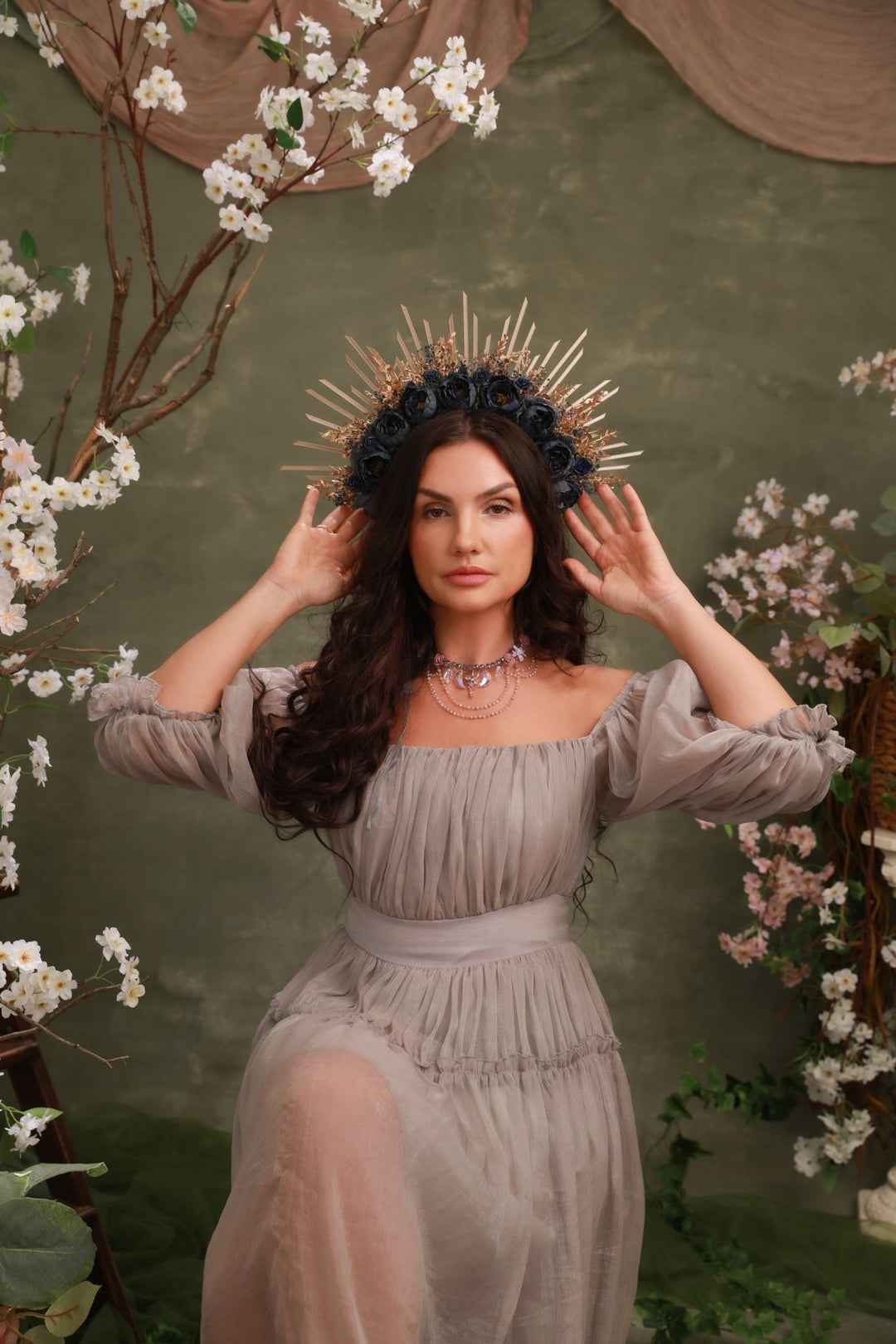 A woman wearing a sheer taupe off-the-shoulder maxi dress and a gold spiked crown, posing in front of a rustic green backdrop decorated with white blossoms.