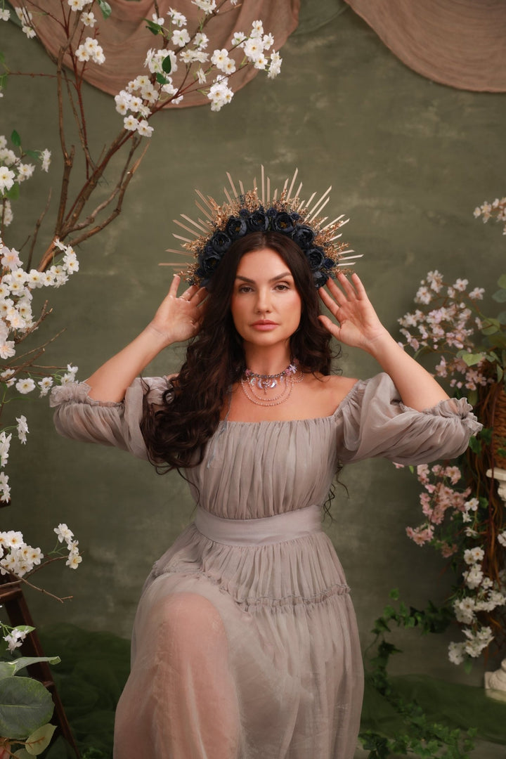 A woman wearing a sheer taupe off-the-shoulder maxi dress and a gold spiked crown, posing in front of a rustic green backdrop decorated with white blossoms.