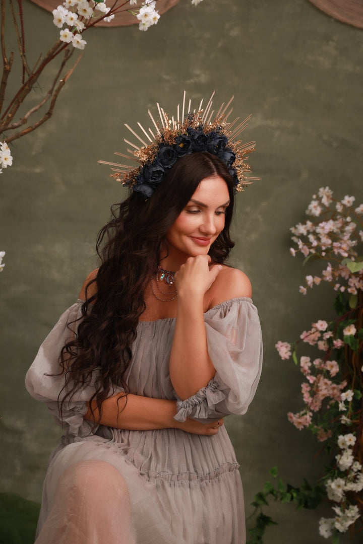 A woman wearing a voluminous off-the-shoulder light gray dress and a decorative crown of dark blue flowers and gold spikes, posed against a textured olive green wall with floral decorations.