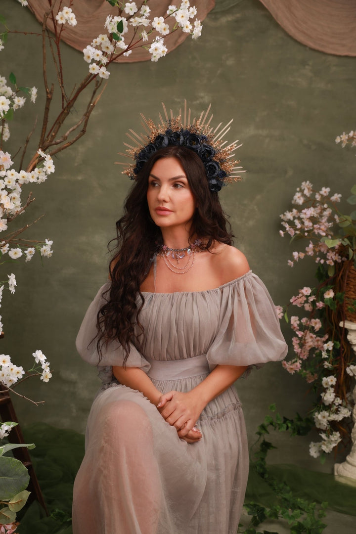 A woman wearing a voluminous off-the-shoulder taupe dress and a gold sunburst crown, posed against a textured olive green backdrop decorated with cherry blossom branches.