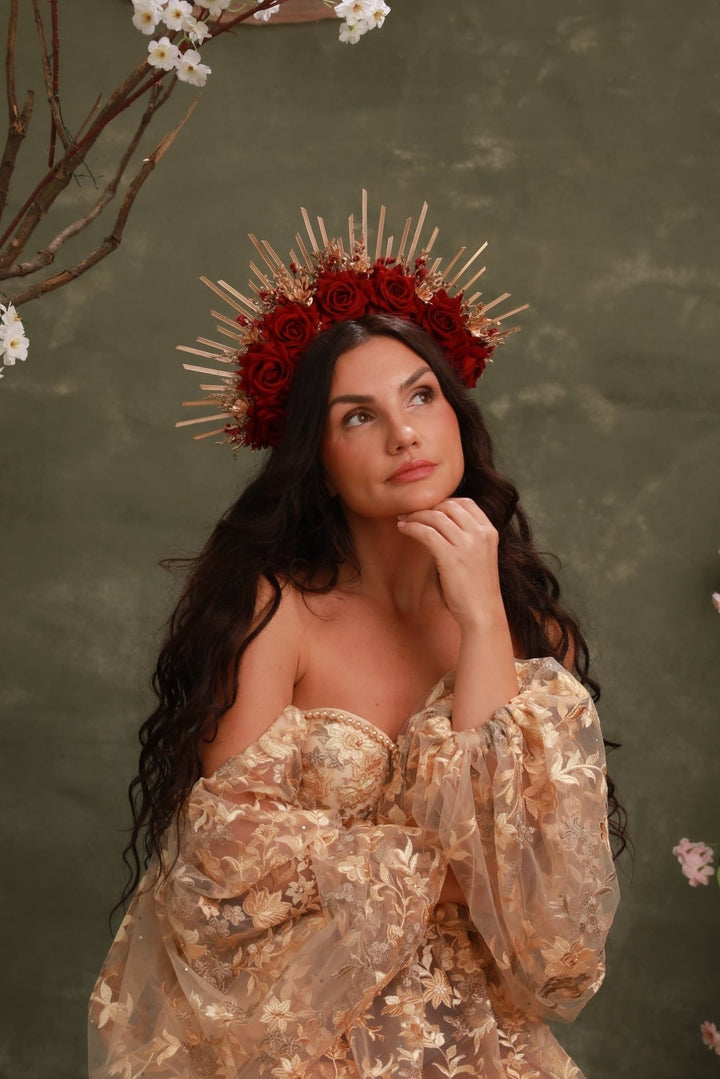A woman wearing a red rose crown with gold spikes and a champagne-gold floral off-the-shoulder dress, posed against a textured olive green background with cherry blossom branches.