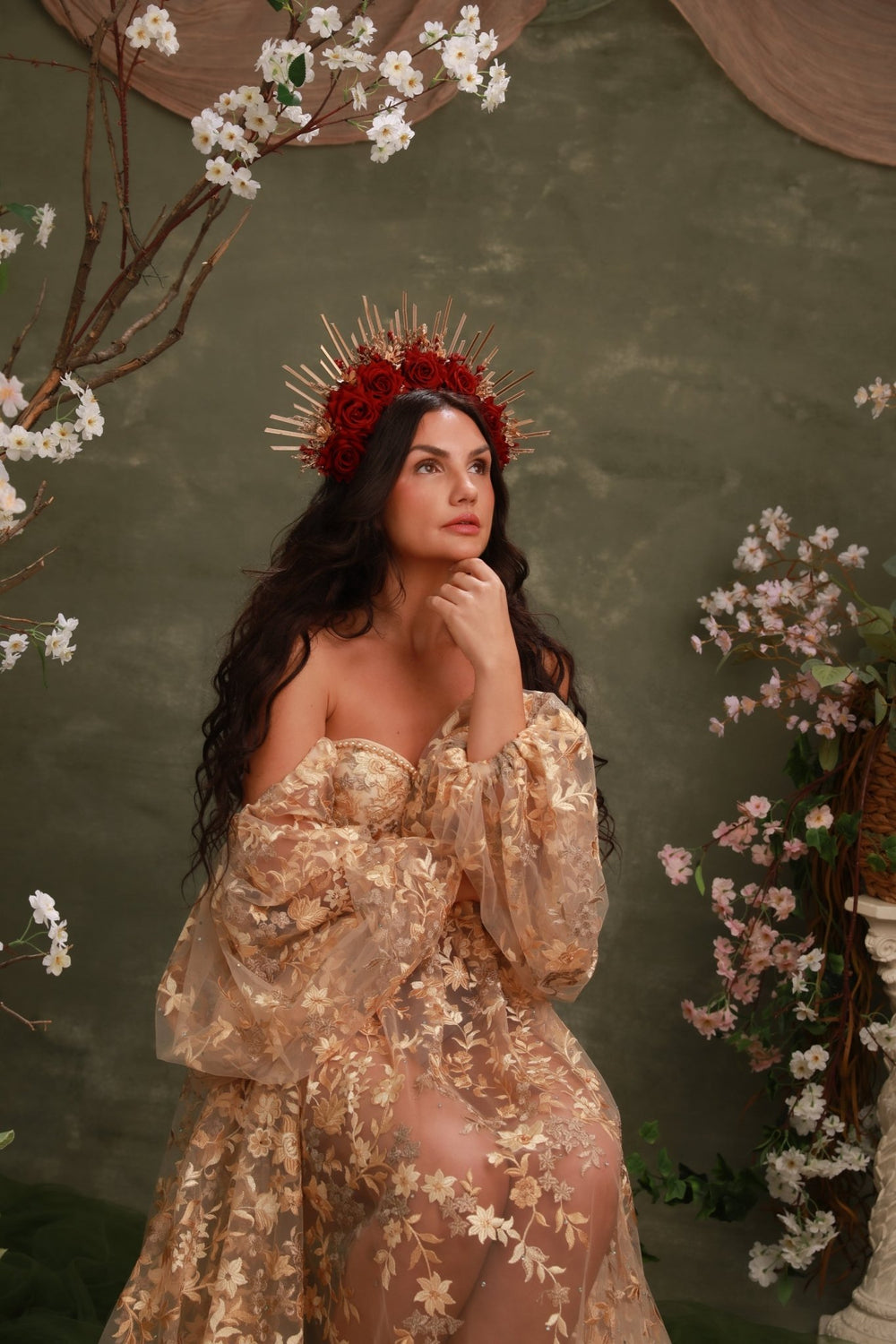 A woman wearing a sheer champagne-gold floral gown with off-the-shoulder puff sleeves and a matching floral crown sits against a textured olive-green backdrop decorated with cherry blossom branches.