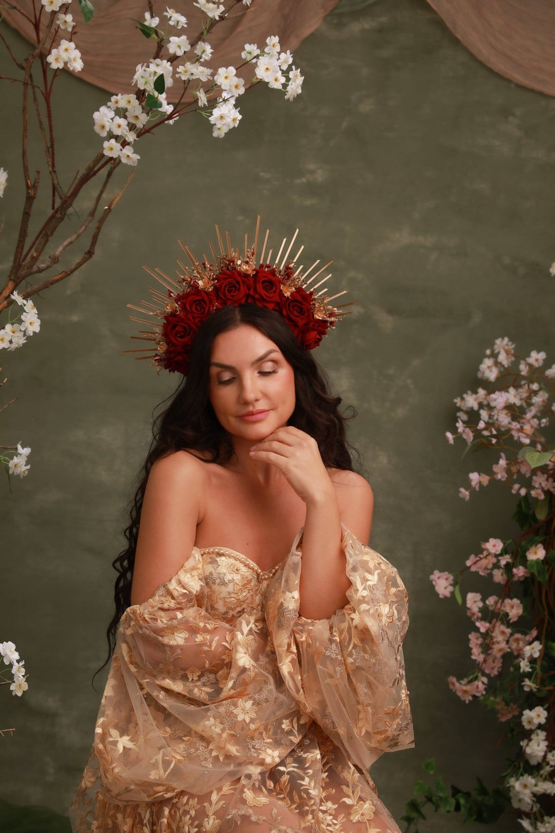 A woman wearing a voluminous champagne-gold off-the-shoulder gown with a matching floral crown of red roses and gold spikes, posed against a textured dark green backdrop with cherry blossom branches.