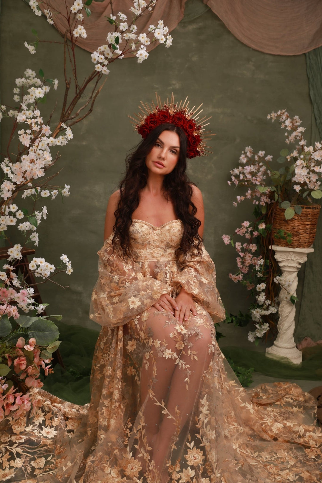 A woman wearing a sheer champagne-gold floral gown with a dramatic train and a red floral crown, posed in front of a dark green backdrop decorated with white blossoms and draped fabric.