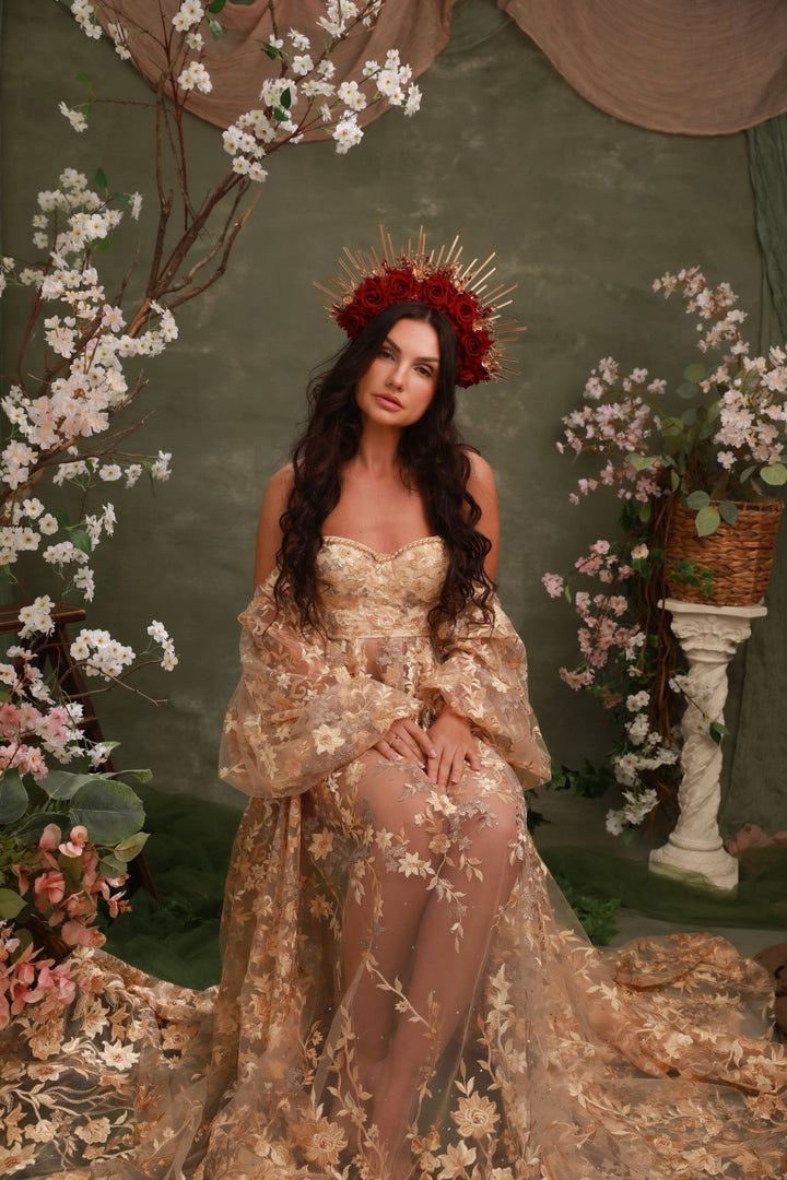A woman wearing a sheer champagne-gold floral gown with a dramatic train and a red floral crown, posed in front of a dark green backdrop decorated with white blossoms and draped fabric.