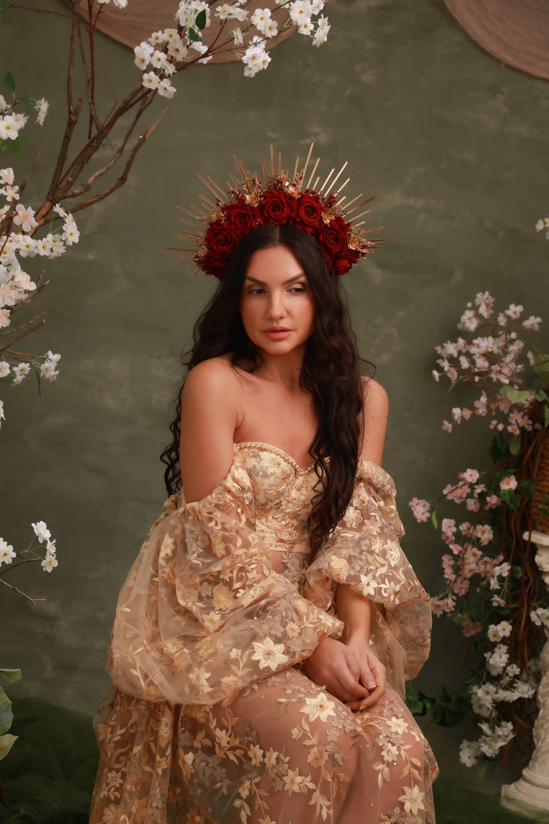 A woman seated in a studio setting wearing a strapless beige floral gown with dramatic ruffled sleeves and a red flower crown with gold spikes. The background is a textured olive-green wall with decorative white and pink blossoms.