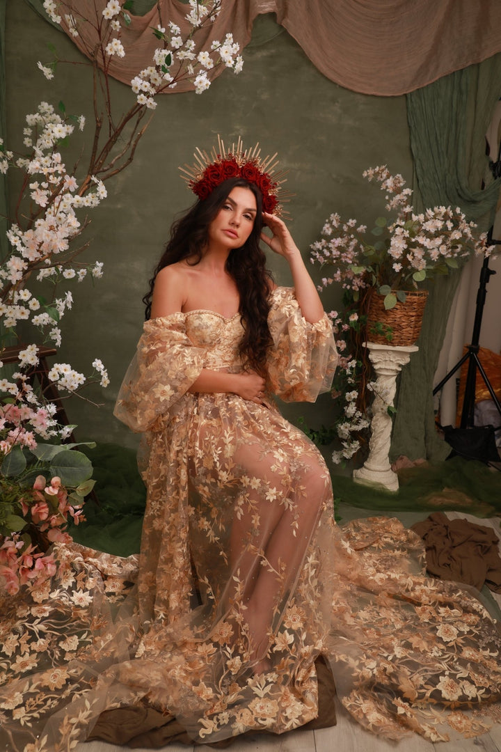 A woman models an off-the-shoulder champagne beige gown with intricate floral embroidery and sheer mesh, styled with a red and gold crown in a themed studio setting with floral arrangements.