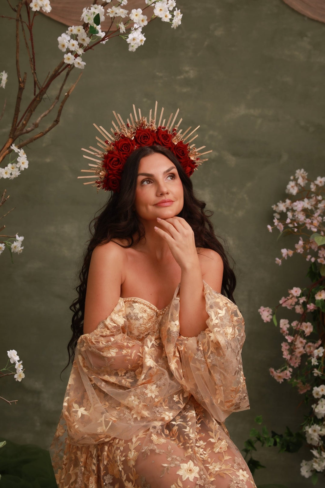 A woman wearing a sheer champagne-gold floral dress and a red rose crown with gold spikes, posed against a textured olive-green backdrop with cherry blossom branches.