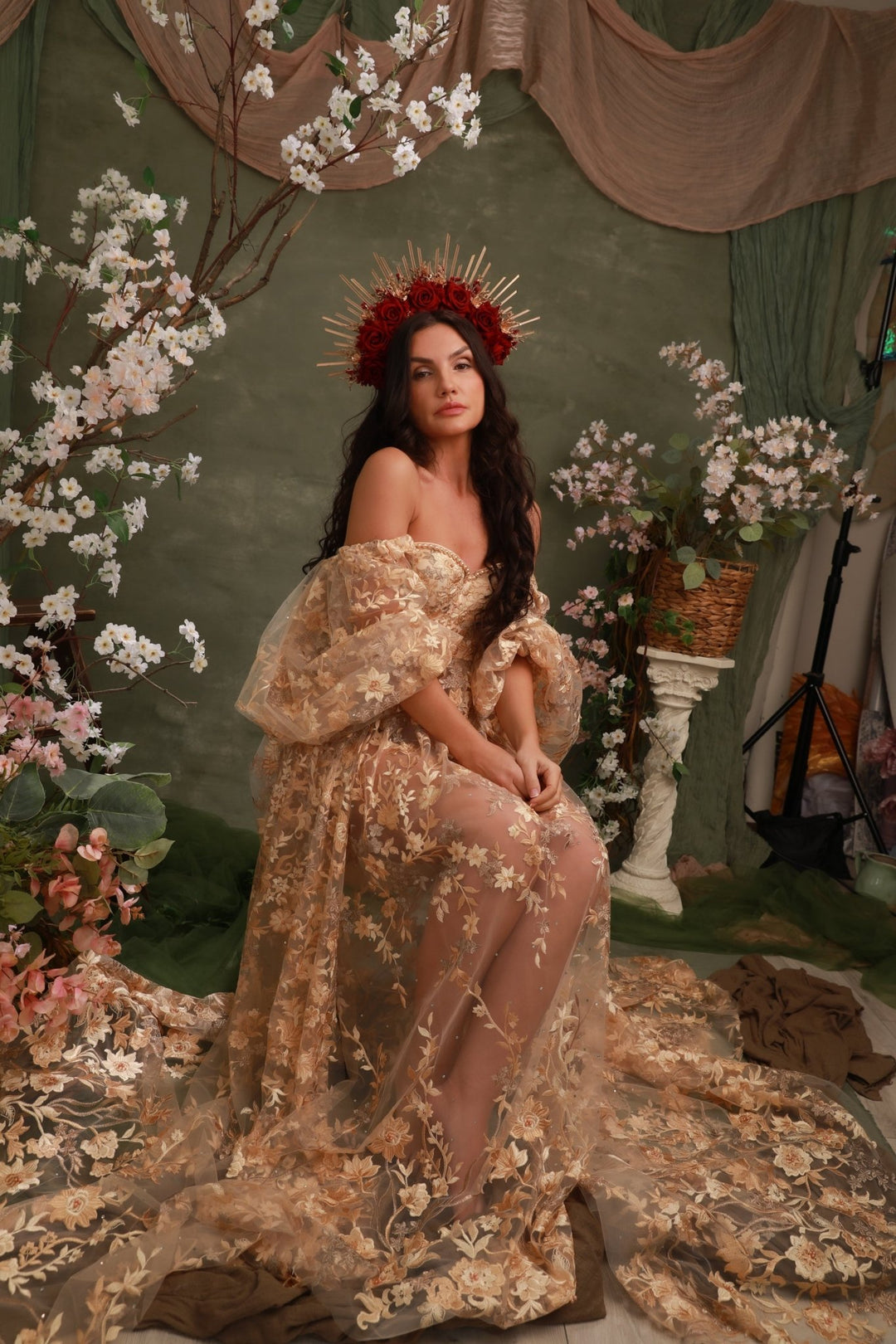 A woman seated in a fantasy-style studio scene wearing an ornate off-the-shoulder beige and gold floral gown with sheer tulle, a red floral crown, and a long train spread across the floor. The set includes draped green fabric, hanging blossoms, and decorative props.