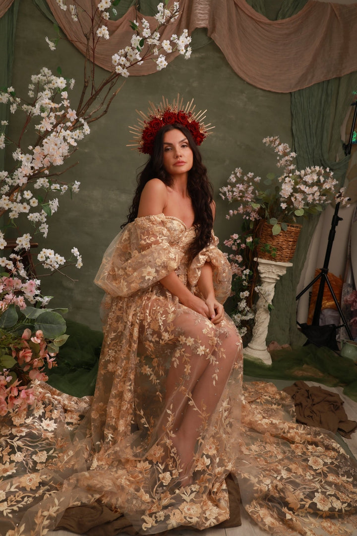 A woman seated in a fantasy-style studio scene wearing an ornate off-the-shoulder beige and gold floral gown with sheer tulle, a red floral crown, and a long train spread across the floor. The set includes draped green fabric, hanging blossoms, and decorative props.
