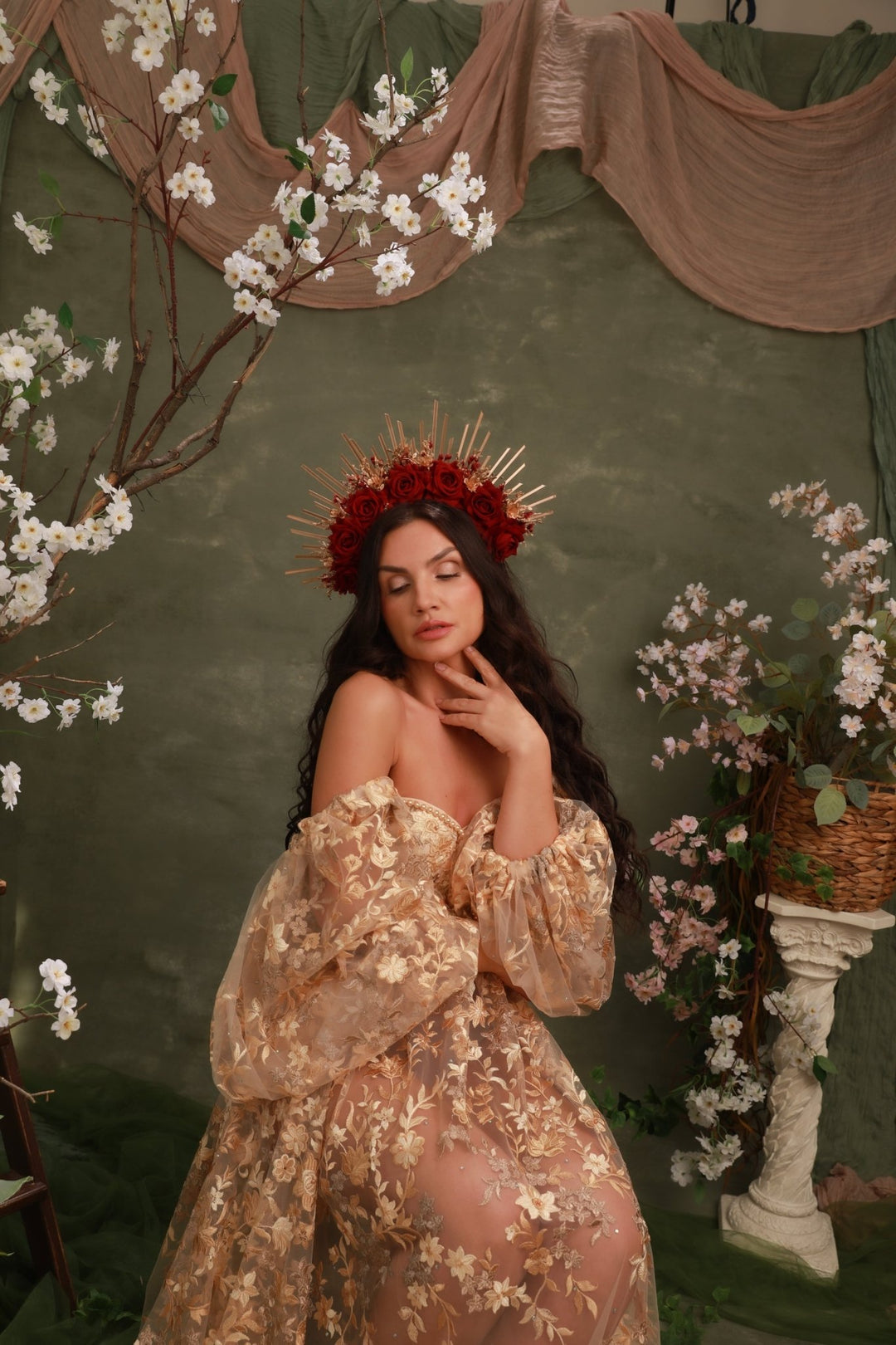 A woman wearing a sheer champagne-gold off-the-shoulder dress with floral embroidery and a red floral crown, posed against a dark green backdrop with draped fabric, white blossoms, and a decorative pedestal.