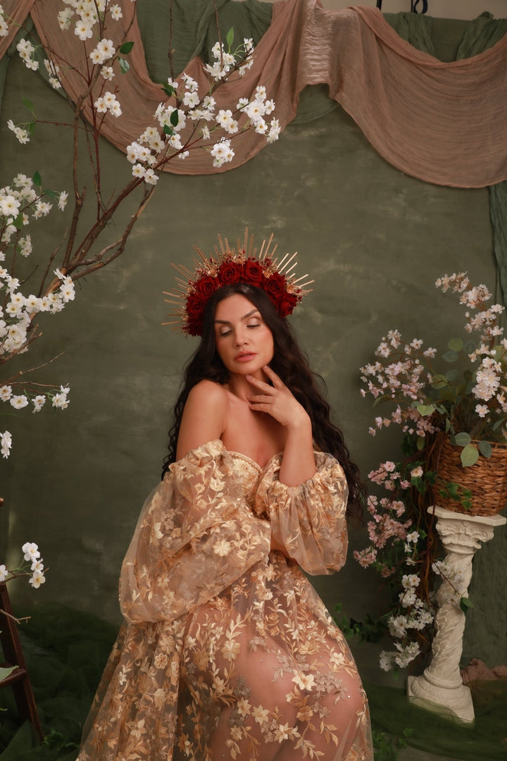 A woman wearing a sheer champagne-gold off-the-shoulder dress with floral embroidery and a red floral crown, posed against a dark green backdrop with draped fabric, white blossoms, and a decorative pedestal.