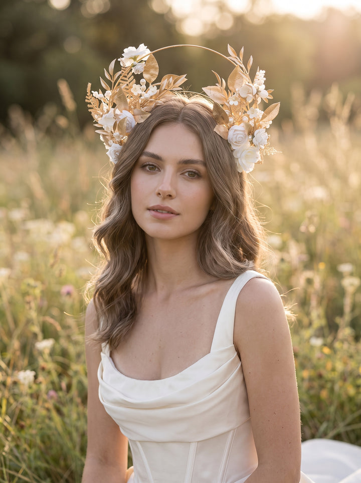 A woman wearing a bohemian-style flower and leaf crown sits in a sunlit grassy field. The crown features white and cream flowers with tan and gold-toned dried botanical elements.