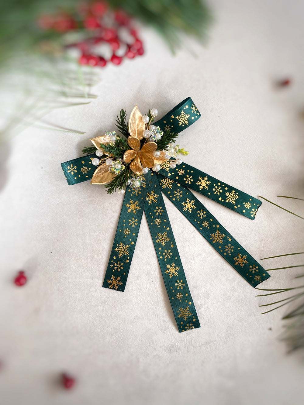 A decorative Christmas bow ornament featuring dark green ribbon with gold snowflake patterns, accented with gold poinsettias, white faux berries, and green pine needles, displayed on a textured light background with festive blurred pine branches and red berries.