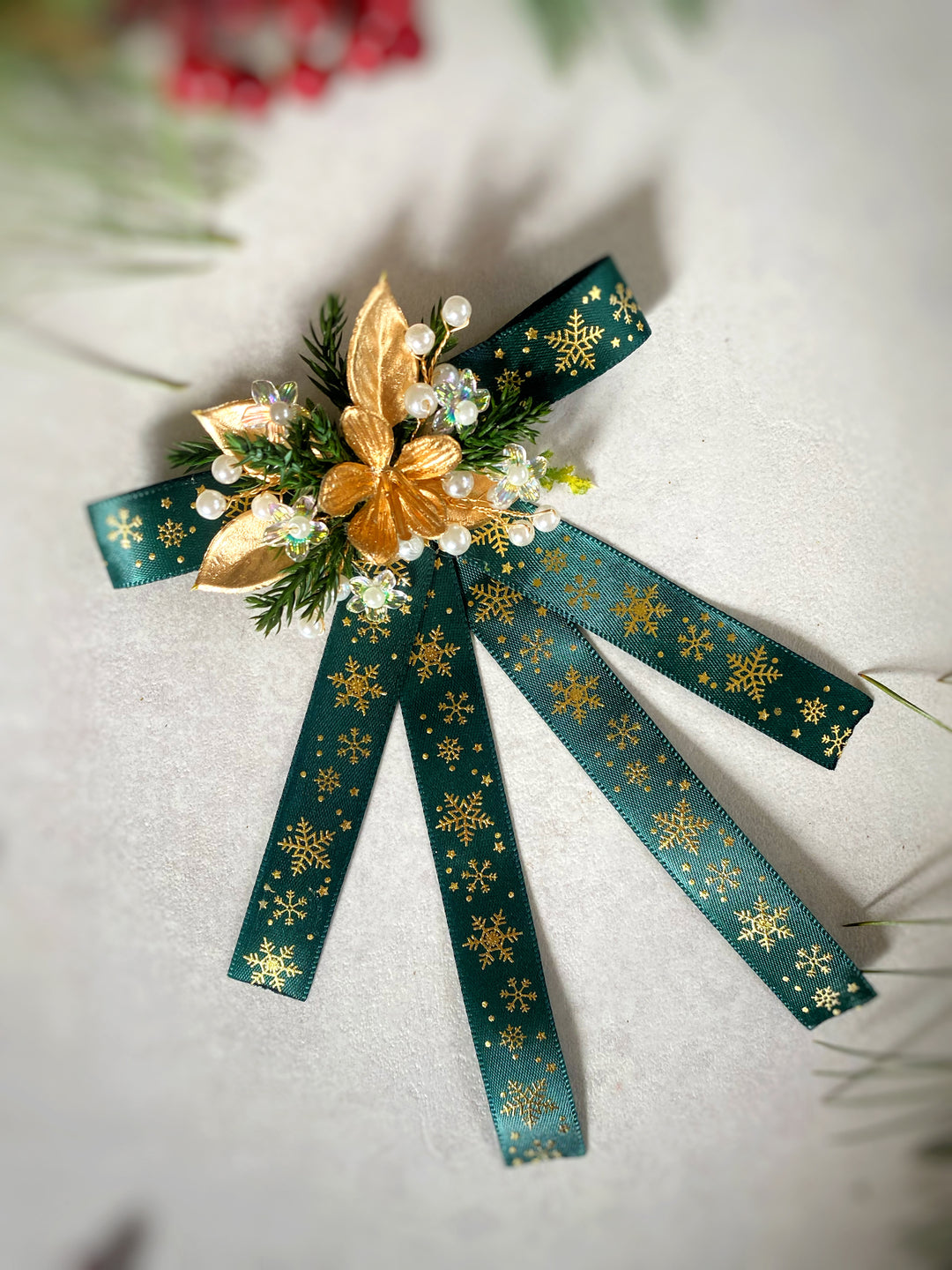 A decorative Christmas ornament or bow featuring dark teal-green ribbon with gold snowflake patterns, a gold poinsettia, green pine needles, and small white faux pearls on a light textured background.