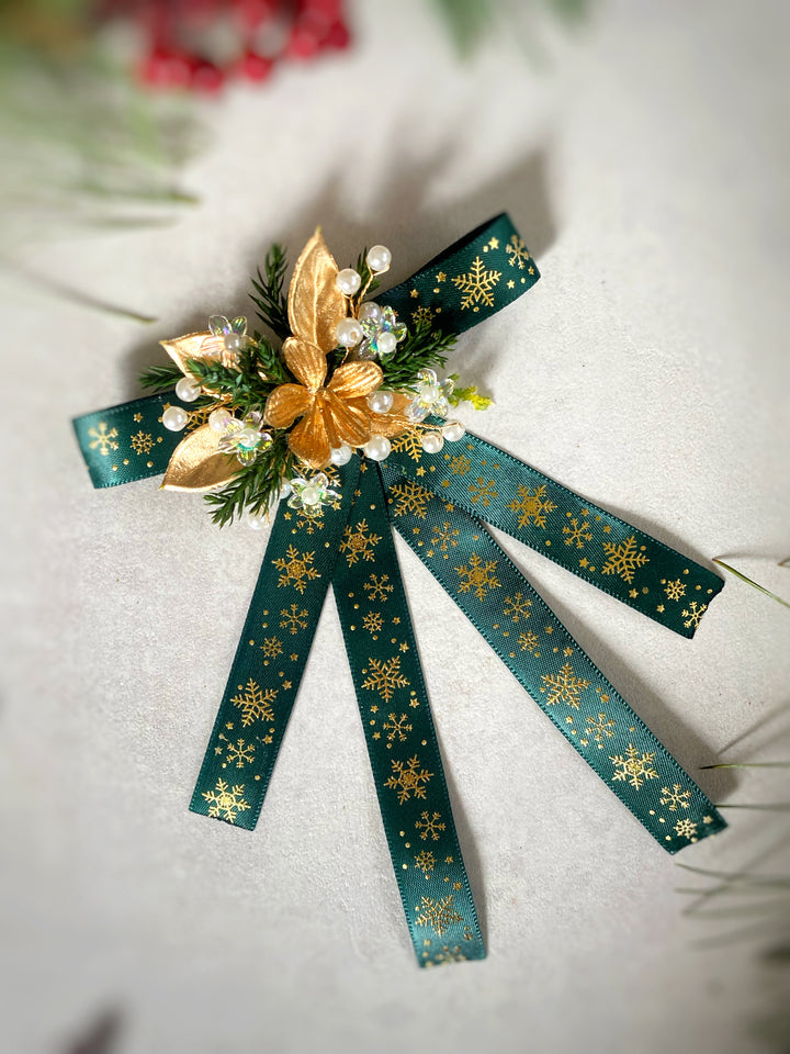 A decorative Christmas ornament or bow featuring dark teal-green ribbon with gold snowflake patterns, a gold poinsettia, green pine needles, and small white faux pearls on a light textured background.
