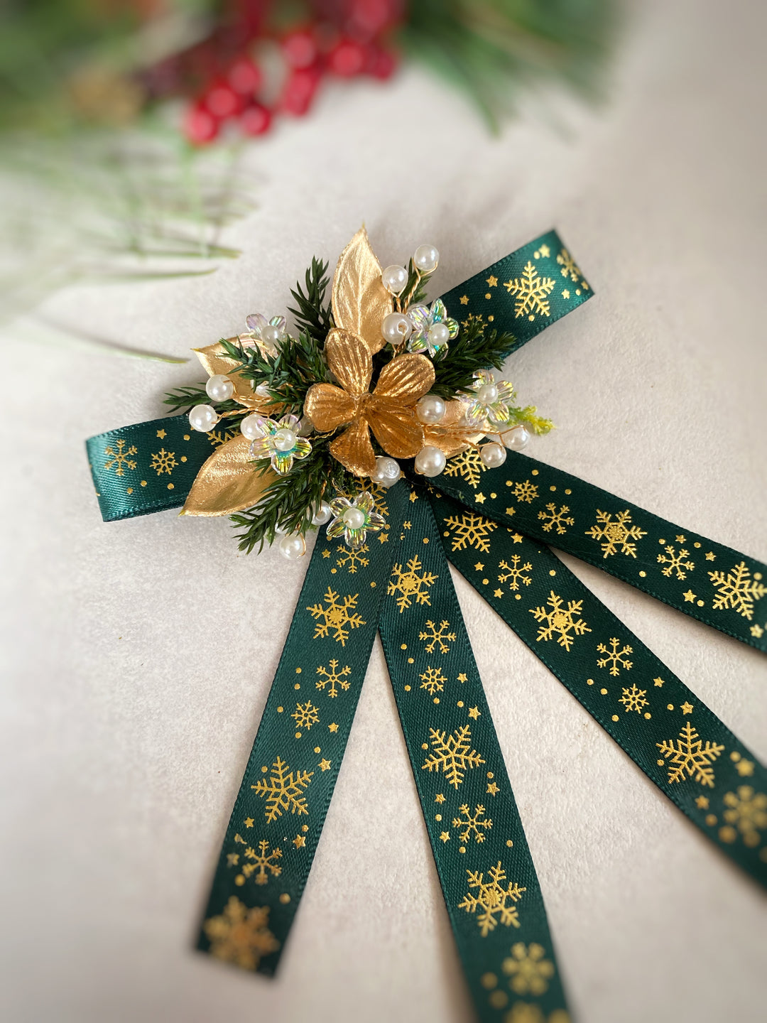 A festive Christmas hair bow featuring dark green ribbon with gold snowflake patterns, decorated with a gold poinsettia, green pine needles, and faux pearls, set against a soft beige background.