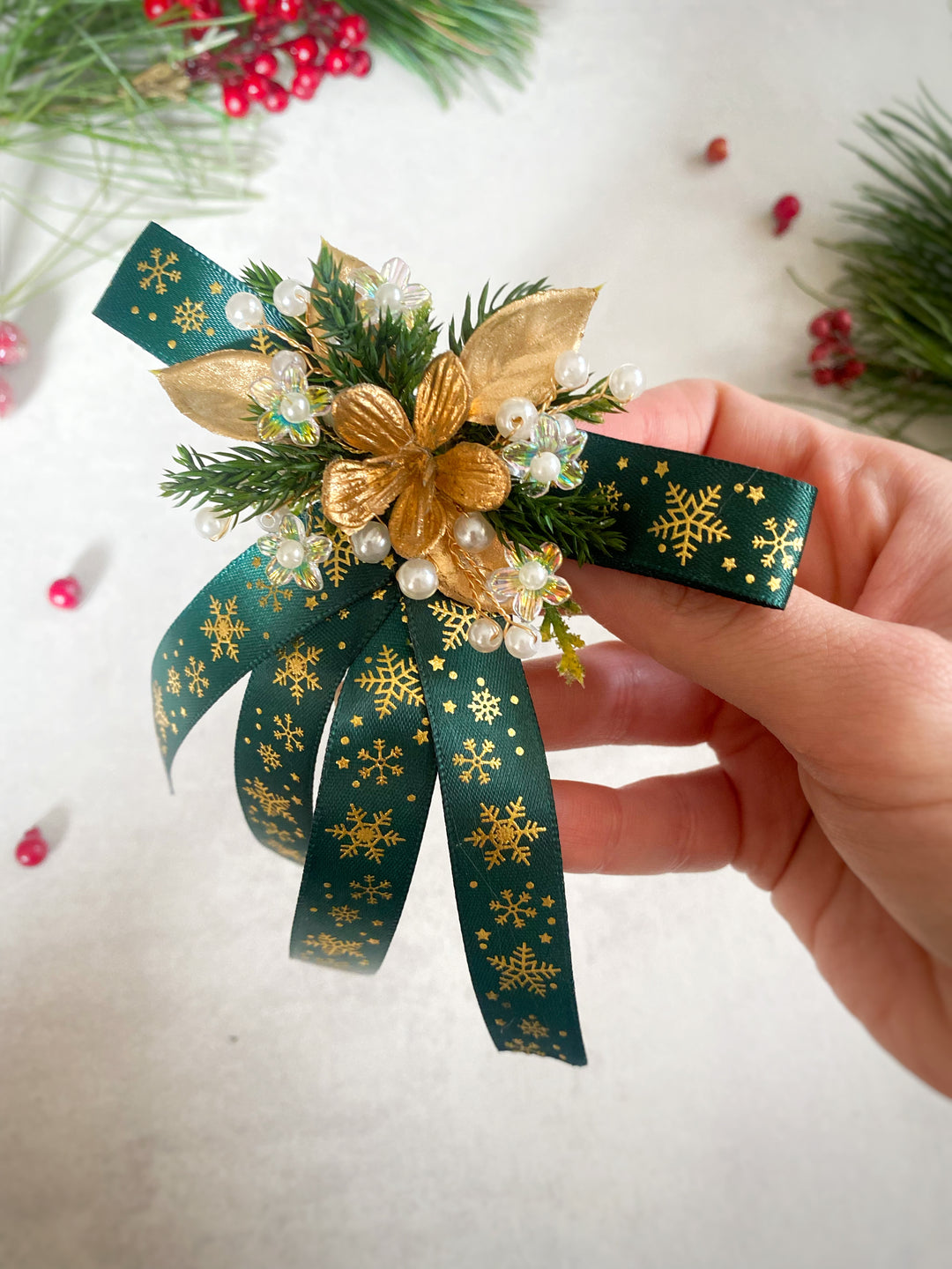 A hand holding a green and gold Christmas hair bow accessory decorated with pine branches, a gold poinsettia flower, white pearl beads, and gold snowflake-patterned ribbon on a light background.