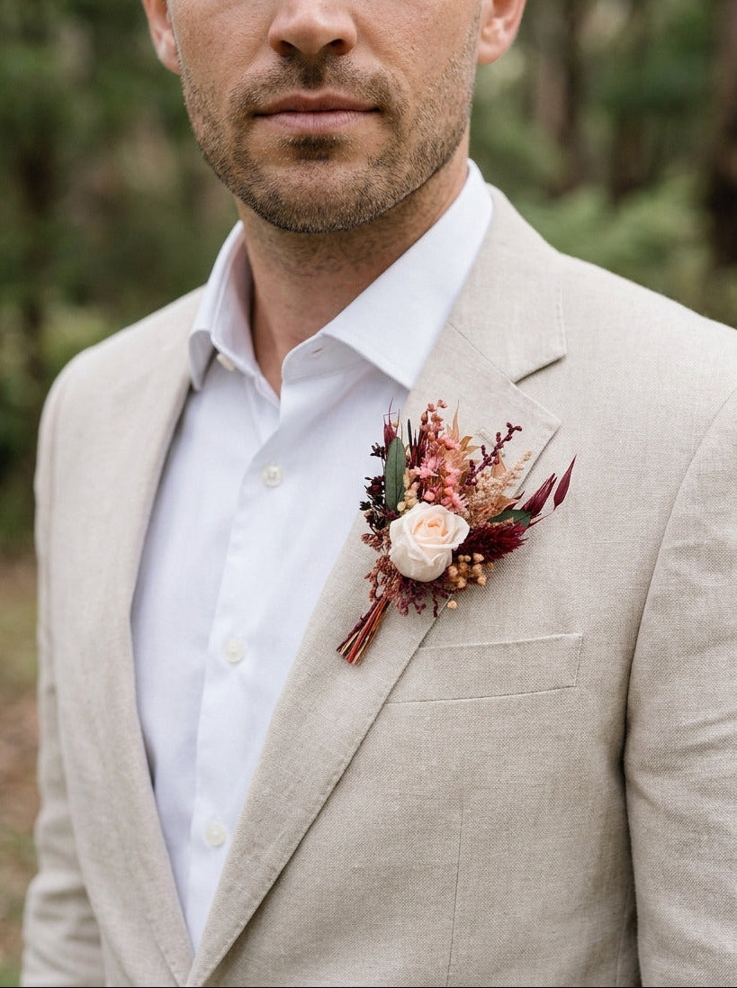 A man wearing a light beige linen suit jacket over a white dress shirt, featuring a rustic floral boutonniere with burgundy, pink, and cream flowers on the chest, set against a blurred forest background.