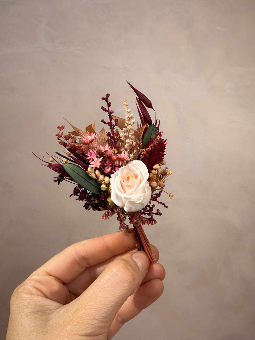 A hand holding a small dried floral boutonniere featuring a pale peach rose, burgundy and pink filler flowers, and green leaves against a neutral textured beige background.