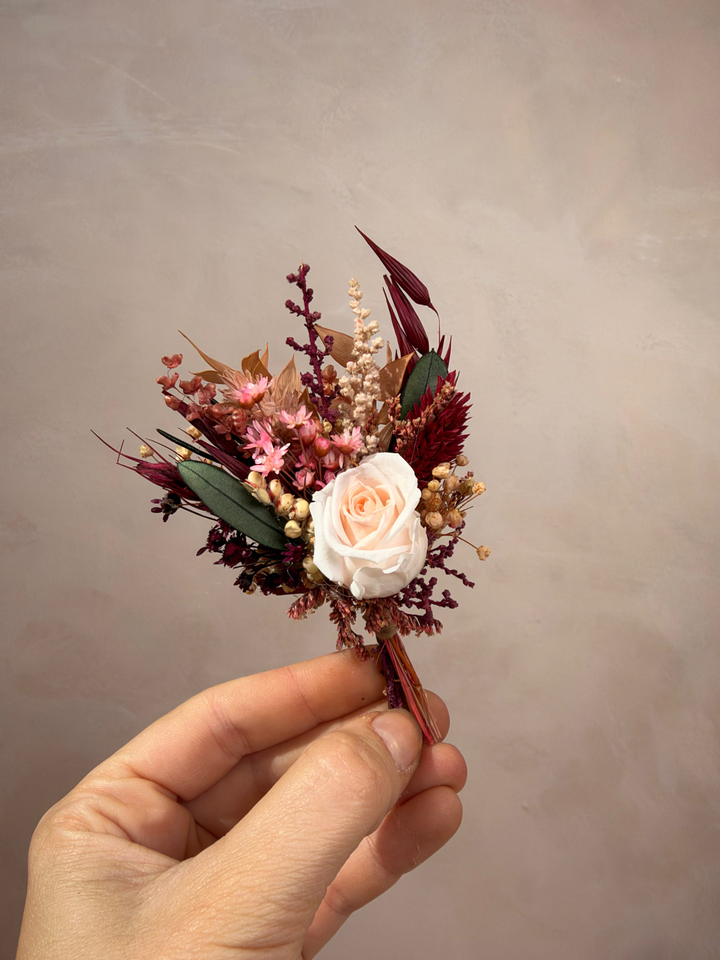 A hand holding a small dried floral boutonniere featuring a pale peach rose, burgundy and pink filler flowers, and green leaves against a neutral textured beige background.