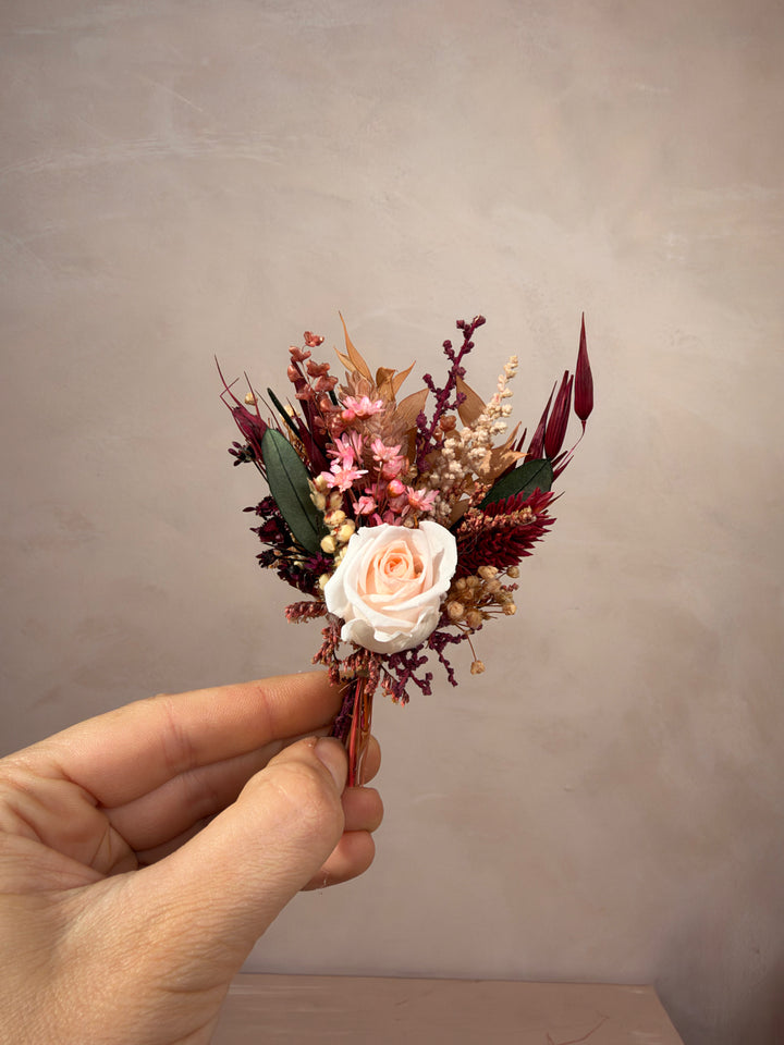 A hand holding a small dried floral boutonniere featuring a pale peach rose, burgundy and pink filler flowers, and green leaves against a neutral textured beige background.