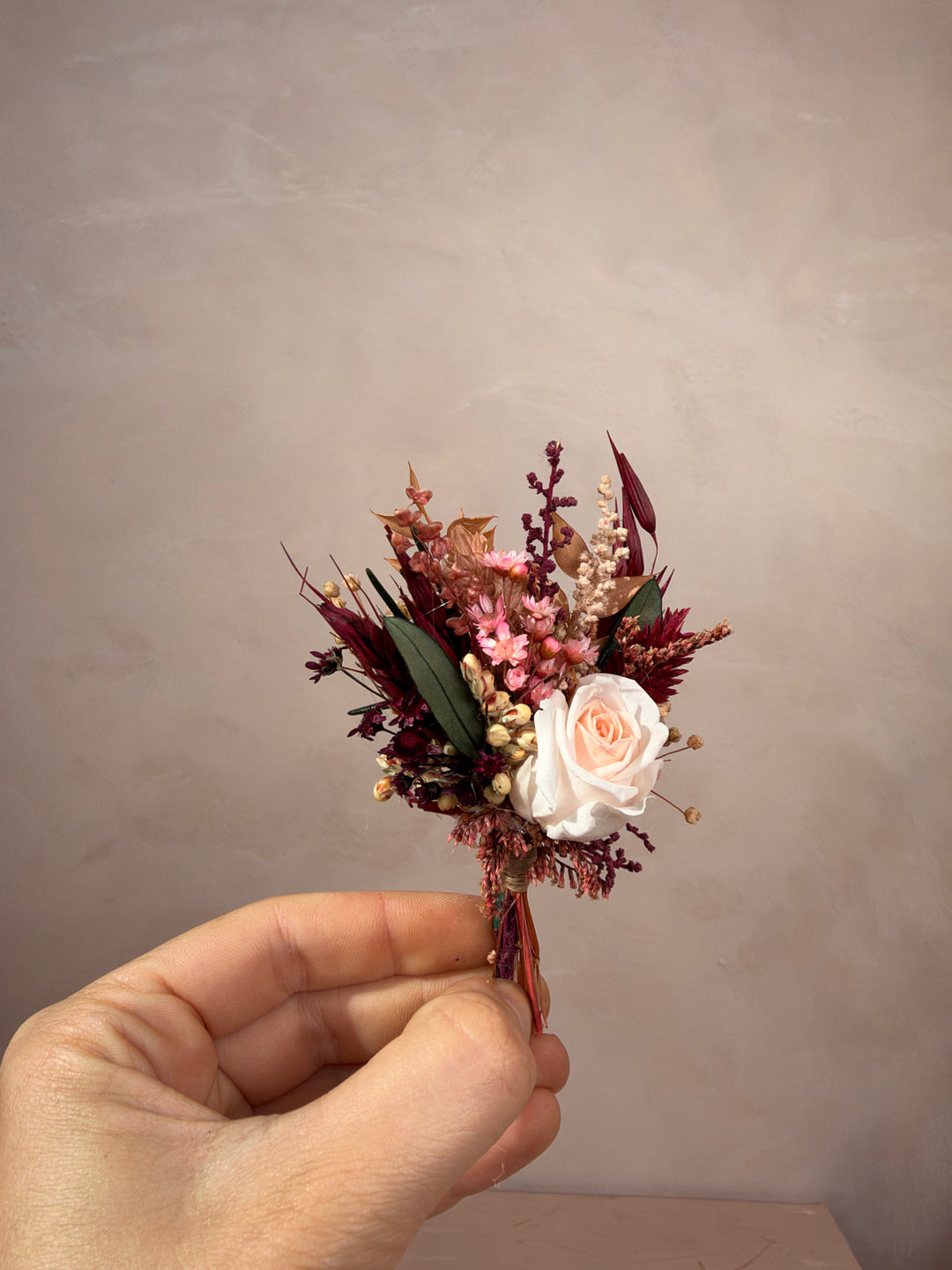 A hand holding a small dried floral boutonniere featuring a pale peach rose, burgundy berries, green leaves, and beige filler stems against a neutral textured beige background.