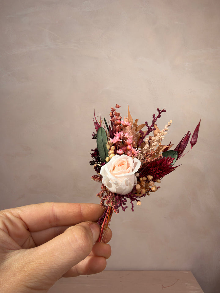 A hand holding a small dried floral boutonniere featuring a pale pink rose, burgundy and pink filler flowers, and green leaves against a neutral textured beige background.