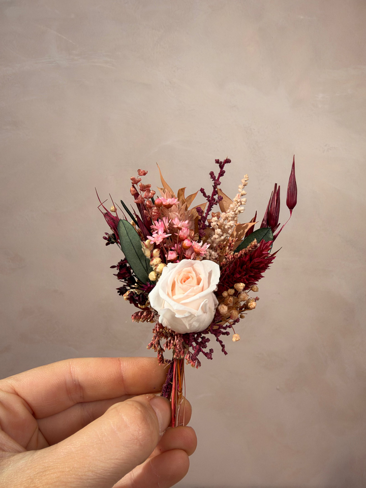 A hand holding a small dried floral boutonniere featuring a pale pink rose, burgundy and pink filler flowers, and green leaves against a neutral textured beige background.
